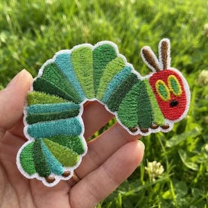 May include: An embroidered patch shaped like a caterpillar with a red head, yellow eyes, and a segmented body featuring various shades of green and teal. The design includes two brown antennae and small brown feet along the bottom edge.