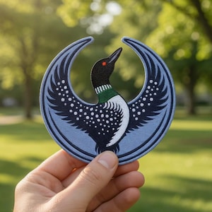Op de afbeelding: Een cirkelvormige patch met een gestileerd ontwerp van een ijsvogel. De patch heeft een lichtblauwe achtergrond met de vleugels van de vogel gespreid. De vogel heeft een zwarte kop, een witte borst en een groene nek. De ijsvogel heeft een rood oog en kijkt omhoog.