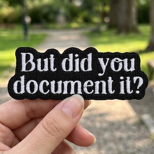 以下が含まれることがあります： 「But did you document it?」と白い刺繍が施された黒いパッチ。パッチは波状の縁取りがあり、ぼやけた屋外の背景の前に保持されています。
