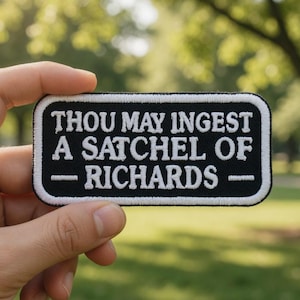 Può includere: Una toppa in bianco e nero con la scritta "THOU MAY INGEST A SATCHEL OF - RICHARDS -". La toppa ha un bordo bianco ed è tenuta davanti a uno sfondo verde sfocato.