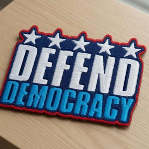 Puede incluir: Parche bordado con las palabras "DEFEND DEMOCRACY" en blanco y azul sobre un fondo azul marino. El parche tiene un borde rojo y estrellas blancas en la parte superior. El parche es rectangular y está sobre una superficie de madera clara.
