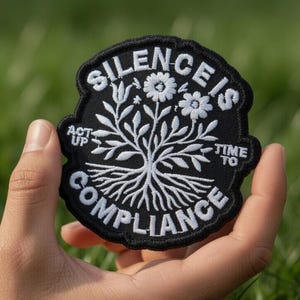 May include: A black and white embroidered patch with the words "SILENCE IS COMPLIANCE" and "ACT UP TIME TO" surrounding a tree design with flowers. The patch is held in a hand against a green grass background.
