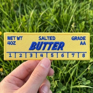 May include: A yellow patch with the word "BUTTER" in blue, and the text "SALTED" and "GRADE AA". The patch also has the text "NET WT 4OZ" and a numbered scale from 1 to 8. The patch is held in front of a grassy background.