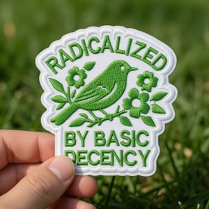 Puede incluir: Parche bordado con un pájaro verde posado en una rama con flores. El parche dice "RADICALIZED BY BASIC DECENCY" en letras blancas sobre un fondo blanco. El parche se sostiene sobre un fondo de hierba verde.