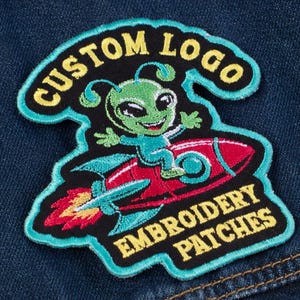 Puede incluir: Parche bordado con un alienígena verde montado en un cohete rojo. El parche tiene el texto "CUSTOM LOGO" y "EMBROIDERY PATCHES" en letras amarillas, delineadas en negro y verde azulado. El parche está sobre un fondo de mezclilla azul.