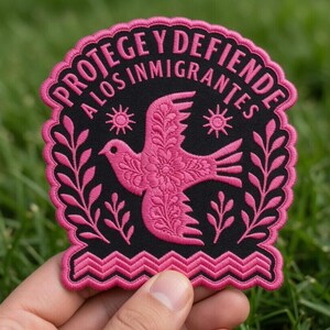 Puede incluir: Un parche bordado en negro y rosa con el texto "PROTEGE Y DEFIENDE A LOS INMIGRANTES" en un diseño curvo. El parche presenta una paloma rosa con detalles florales, rodeada de hojas y soles rosas.