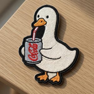 Puede incluir: Parche bordado con un ganso blanco sosteniendo una lata de refresco con una pajita roja. La lata es gris con la palabra "CoCola" en rojo. El ganso tiene pico y patas naranjas, con un contorno negro.