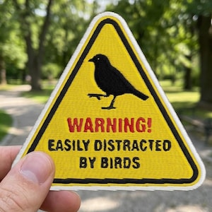 May include: Yellow triangular patch featuring a black bird silhouette and the text "WARNING! EASILY DISTRACTED BY BIRDS" in red and black. The patch has a black border. The background is a blurred outdoor scene.