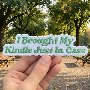 May include: A white patch with green embroidered text that reads "I Brought My Kindle Just In Case." The patch has a scalloped edge and is held in front of a park scene with trees and benches.