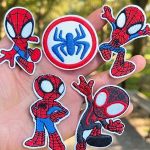 May include: Five embroidered patches featuring superhero characters and a spider emblem. Three patches show a red and blue character in various poses, one shows a black and red character in a dynamic pose, and one is a circular patch with a blue spider.