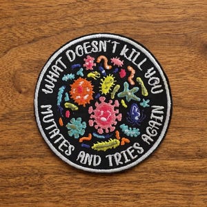 以下が含まれることがあります： 「WHAT DOESN'T KILL YOU MUTATES AND TRIES AGAIN」の白い文字が入った、黒い丸い刺繍パッチ。パッチには、さまざまな微生物のカラフルな刺繍画像が施されています。