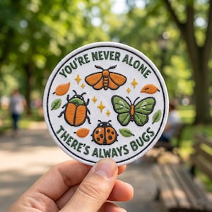 May include: A circular white patch with the text "YOU'RE NEVER ALONE THERE'S ALWAYS BUGS" in green. The patch features orange and green illustrations of insects, including a moth, beetle, butterfly, and ladybug. The patch is held in a hand.