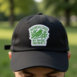 Puede incluir: Gorra de b&eacute;isbol negra con un parche blanco con un p&aacute;jaro verde, flores y el texto "RADICALIZED BY BASIC DECENCY". La gorra tiene una visera curva y una correa ajustable.