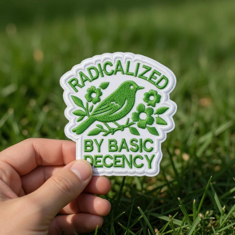 Puede incluir: Parche bordado con un p&aacute;jaro verde posado en una rama con flores. El parche dice "RADICALIZED BY BASIC DECENCY" en letras blancas sobre un fondo blanco. El parche se sostiene sobre un fondo de hierba verde.