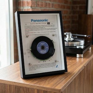 以下が含まれることがあります： 黒いフレームに入ったパナソニックSL-CT700ポータブルCDプレーヤー。フレームには黒いレコード盤と回路基板が入っています。フレームには「パナソニック」の文字と製品仕様が表示されています。フレームは木製のキャビネットに置かれています。