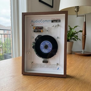 May include: A framed AudioComm CD player, model CDP-8502-W, with a black vinyl record. The frame is brown, and the background is white. The text "From Japan" is visible. Two batteries are also visible.