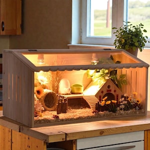 Peut inclure: Cage en bois pour hamster avec un dessus en verre. La cage comprend une petite maison en bois, une rampe et des fleurs décoratives. L'intérieur est éclairé par un éclairage chaleureux, créant un environnement confortable pour les petits animaux.