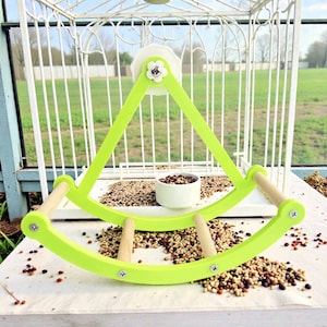 May include: A lime green bird perch with wooden dowel handles and a small white ceramic bowl. The perch is attached to a white birdcage. Birdseed is scattered around the perch.