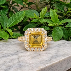May include: A gold ring featuring a large, square-cut yellow gemstone at the centre, surrounded by a halo of small, clear, round-cut stones. The band is also encrusted with small, clear, round-cut stones along the sides of the setting.