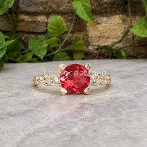 Op de afbeelding: Een gouden ring met een grote, ronde, felrode middensteen die door vier klauwen wordt vastgehouden. De ringband is gemaakt van getextureerd goud en is bezet met een reeks kleine, heldere, rond geslepen stenen langs beide zijden.