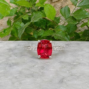 Può includere: Un anello color argento con una grande pietra preziosa rosso vivo a taglio cuscino rettangolare come elemento centrale, affiancato da una fascia incastonata con piccole pietre chiare e scintillanti sui lati, su una superficie di pietra neutra.