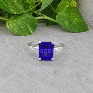 May include: A silver ring featuring a large, rectangular, deep blue gemstone set in a four-prong mounting. The band is smooth and polished, showcasing a clean, metallic finish that contrasts with the vibrant, saturated blue of the centre stone.