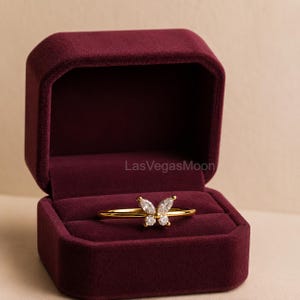 May include: A gold-colored ring with a butterfly design, set with clear gemstones. The ring is displayed inside a burgundy velvet ring box. The text "LasVegasMoon" is visible on the box.