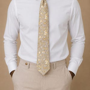 May include: A man in a white shirt, beige trousers, and a patterned tie. The tie has a beige base with a floral pattern in yellow and light brown. The man's hands are in his pockets. The tie's pattern is visible.