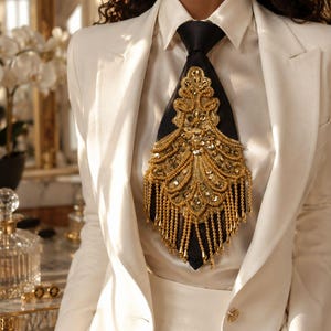 May include: A black necktie featuring an elaborate gold-toned embroidered appliqué with sequins, beads, and dangling fringe accents. The tie is worn over a white satin button-down shirt and paired with a white tailored blazer with a single gold button.