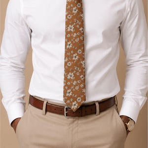 May include: A brown floral tie is the focal point of this image. The tie is worn with a white dress shirt, khaki pants, and a brown leather belt. A gold watch is visible on the wrist. The tie features a repeating pattern of small white flowers.