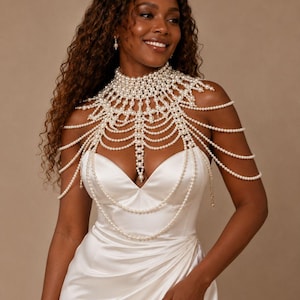 May include: A white pearl shoulder necklace featuring a high collar design with multiple draped strands of pearls that extend across the chest and shoulders. The accessory is constructed from numerous small, round, white pearls arranged in a complex, layered pattern that covers the upper torso and neckline.