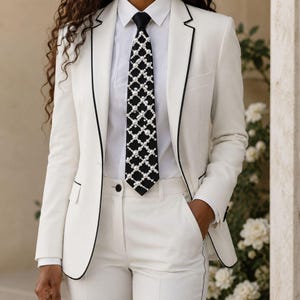 May include: A white blazer and matching trousers set with thin black piping along the lapels and pockets. The outfit includes a crisp white button-down shirt and a black necktie featuring a white lattice pattern embellished with small white pearls.