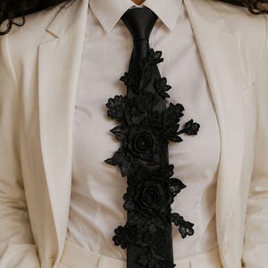 May include: A black necktie featuring an intricate black floral lace appliqué that extends down the center. The tie is worn over a crisp white button-down shirt and a white blazer, creating a high-contrast look with detailed fabric textures.