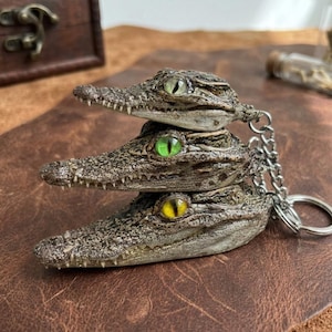 May include: A keyring featuring three stacked crocodile head replicas. Each head has a textured, mottled brown and grey surface with visible teeth and a distinct eye: one pale green, one bright green, and one yellow, all attached to a metal chain.
