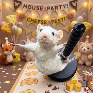 May include: A white mouse figurine mounted on a black circular base, holding a black ridged rod. The background features a banner with the text MOUSE PARTY and CHEESE FEST, surrounded by small plush animals, cheese-shaped decorations, and gifts.