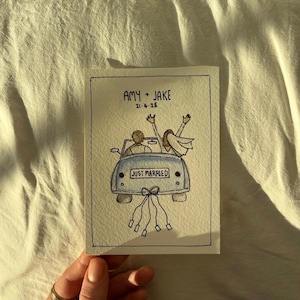 May include: A watercolour illustration of a car with a newly married couple inside, with the words "JUST MARRIED" on the back. The card is labelled "AMY + JAKE 21.6.25". The car has ribbons tied to the back.