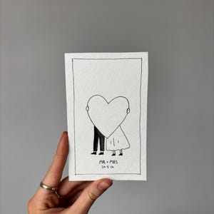 May include: A black and white illustration of a couple embracing behind a heart shape. The artwork is on white paper, with the text "MR + MRS 24-5-24" below. The drawing is framed with a black border and held by a hand.