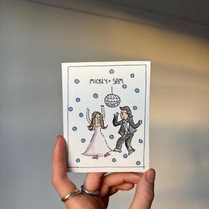 May include: A watercolour illustration of a couple dancing under a disco ball. The artwork features the text "MICKEY + SAM" above the couple, surrounded by blue dots. The woman wears a pink dress, and the man is in a tuxedo.