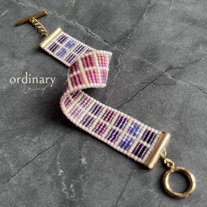 May include: A beaded bracelet with a plaid pattern in shades of purple, pink, and white. The bracelet has gold-coloured metal end caps and a toggle clasp. The text "ordinary mind" is in the image.