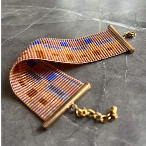 May include: A woven bracelet with a plaid pattern in shades of orange, pink, and blue. The bracelet has gold-colored metal bars at each end and a gold chain clasp. The bracelet is approximately 1.5 inches wide.