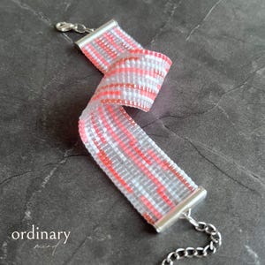 May include: A woven bracelet with a pattern of pink, white, and gray beads. The bracelet has silver-colored end caps and a chain clasp. The word "ordinary mind" is printed in cursive on the bottom left.