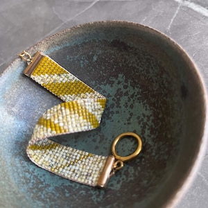 May include: A beaded bracelet with a gold clasp and accents. The bracelet features a pattern of yellow and white beads. The bracelet is displayed in a teal-colored bowl.