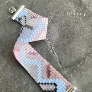 Könnte beinhalten: Ein gewebtes Perlenarmband in Rosa, Blau und Schwarz. Das Armband hat einen silbernen Verschluss und einen kleinen silbernen Anhänger. Das Wort "ordinary" ist im Hintergrund aufgedruckt.