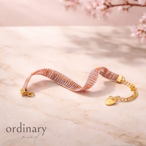 Pode incluir: Uma pulseira tecida com um padrão rosa, laranja e roxo. A pulseira tem um fecho e corrente dourados com um pingente em forma de coração. As palavras "ordinary mind studio" estão na parte inferior da imagem.