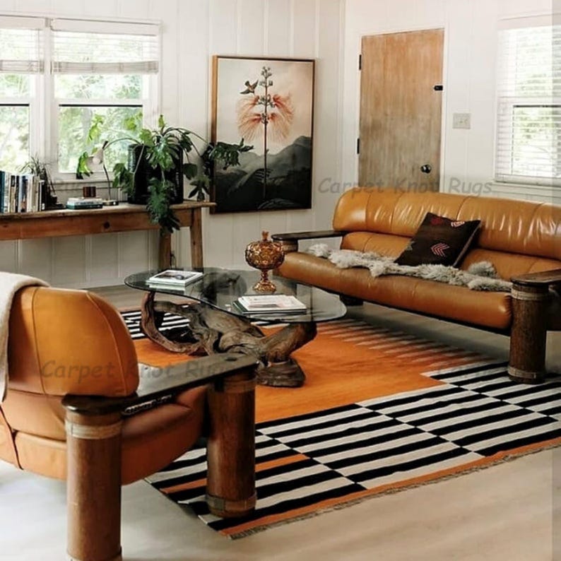 Op de afbeelding: Een woonkamer met een bruine leren bank en fauteuil, een glazen salontafel en een oranje en zwart patroon tapijt. Een schilderij hangt aan de muur en een houten tafel bevat planten en boeken. De kamer heeft een warme, uitnodigende sfeer.