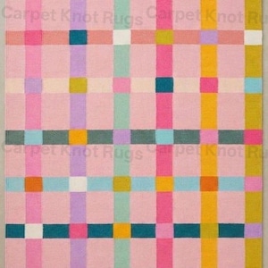 May include: A rectangular rug with a pink background and a colorful plaid pattern. The plaid design features vertical and horizontal stripes in shades of pink, orange, yellow, teal, purple, and white. The rug is a decorative home accent.