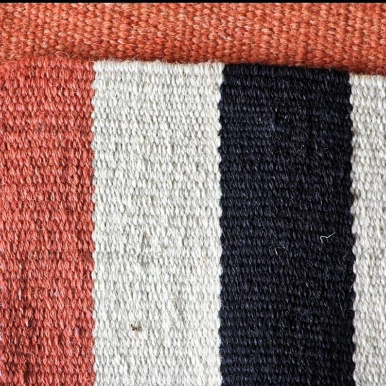 Op de afbeelding: Close-up van een geweven textiel met een streepjespatroon. Het ontwerp heeft verticale strepen in koraal, wit en zwart. Een effen band van oranjerood materiaal bevindt zich bovenaan en voegt een contrasterend element toe aan het ontwerp.