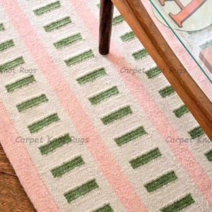 May include: A rectangular rug featuring a repeating pattern of green rectangles and pink stripes on a cream backdrop. The rug is partially visible, with a wooden table leg and part of a glass table top in the frame. The words "Carpet Knots Rugs" are printed on the rug.
