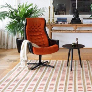 May include: An orange upholstered armchair with a black frame and base. A small black side table with three legs sits on a striped rug. A potted plant and decorative items are in the background.