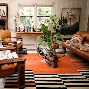 Op de afbeelding: Een woonkamer met een feloranje tapijt met zwart-witte geometrische patronen. De kamer omvat leren stoelen en een bank, een glazen salontafel en een houten boekenplank. Een groot schilderij en planten dragen bij aan de inrichting.